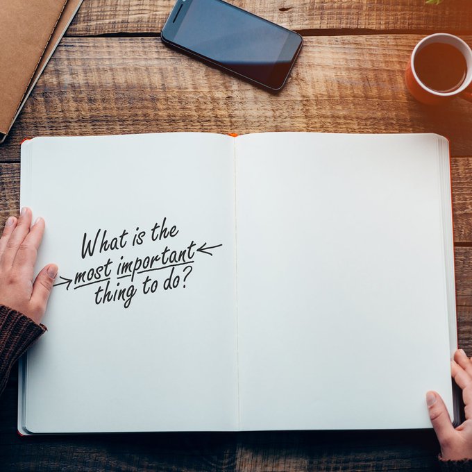 Two hands holding an open journal on a wooden desk. One page of the journal has the handwritten words, What is the most important thing to do?